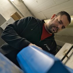 In einem Werkstattumfeld arbeitet ein Mann mit einer blauen Maschine, während er aufmerksam auf seine Aufgabe schaut.