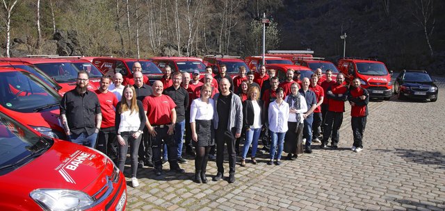 Eine Gruppe von Mitarbeitern steht lächelnd vor einer Reihe roter Fahrzeuge auf einem gepflasterten Platz in der Natur.