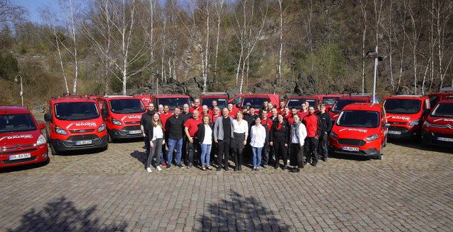 Die fröhliche Gruppe von Angestellten zeigt Teamgeist, während sie vor einer Reihe von roten Transportern auf einem gepflasterten Platz steht.