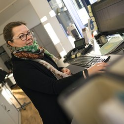In einem modernen Büro lächelt eine Frau, während sie an ihrem Schreibtisch mit einem Computer und Telefon arbeitet.