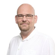 Ein Mann mit Brille und einem kurzen Bart lächelt freundlich vor einem hellen, einfarbigen Hintergrund.