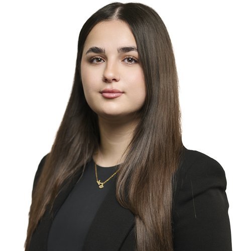 Eine junge Frau mit langen, glatten Haaren und einem selbstbewussten Ausdruck trägt einen schwarzen Blazer und eine goldene Kette.