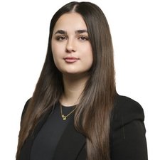 Eine junge Frau mit langen, glatten Haaren und einem selbstbewussten Ausdruck trägt einen schwarzen Blazer und eine goldene Kette.