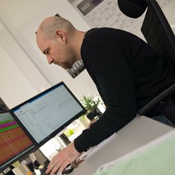 In einem modernen Büro arbeitet ein Mann an zwei Bildschirmen, umgeben von Bürobedarf und einem Kalender im Hintergrund.