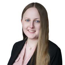 Die Portraitaufnahme zeigt eine Frau mit langen Haaren, die in einem formellen Blazer und einem blassen Oberteil gekleidet ist.