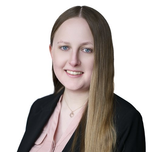 Die Portraitaufnahme zeigt eine Frau mit langen Haaren, die in einem formellen Blazer und einem blassen Oberteil gekleidet ist.