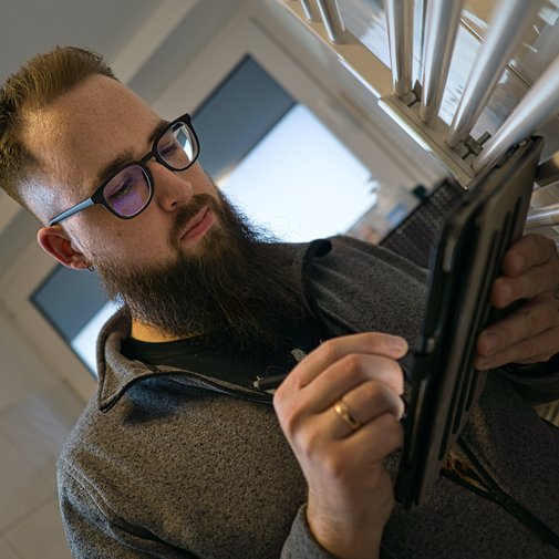 In einem modernen Raum betrachtet ein bärtiger Mann mit Brille sein Tablet, umgeben von einem Heizkörper.